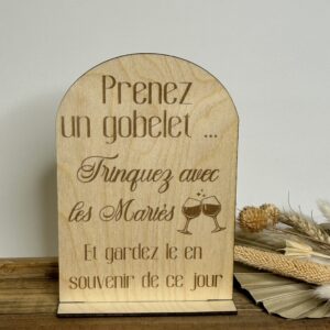 Panneau en bois gravé pour mariage avec inscription “Prenez un gobelet, trinquez avec les mariés” posé sur socle.