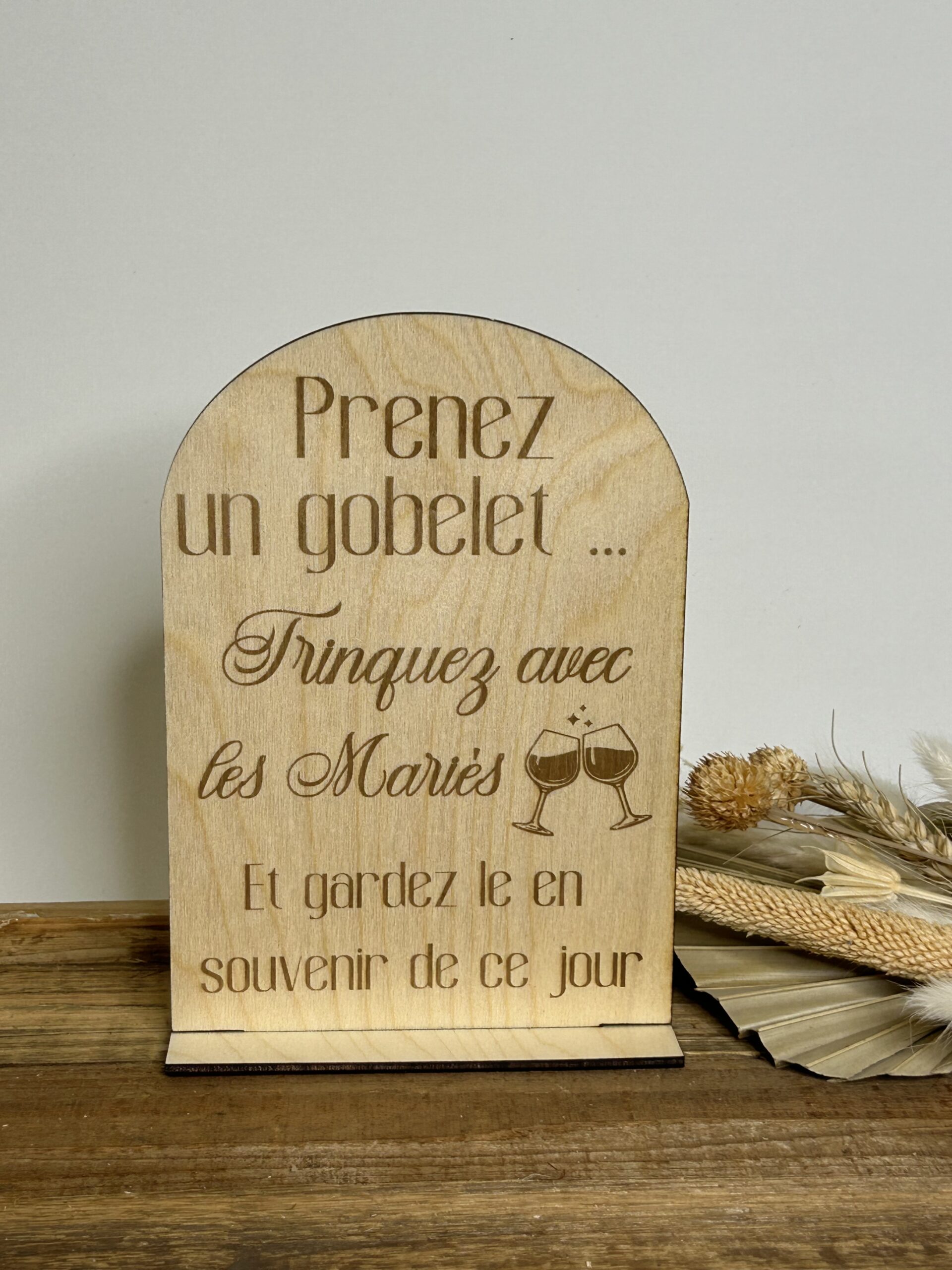 Panneau en bois gravé pour mariage avec inscription “Prenez un gobelet, trinquez avec les mariés” posé sur socle.