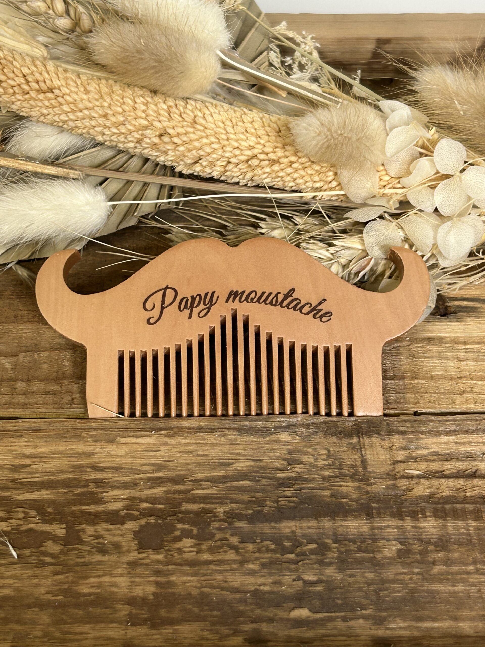 Peigne à moustache et barbe en bois gravé pour papa et papy posé sur fond naturel en bois avec fleurs séchées.